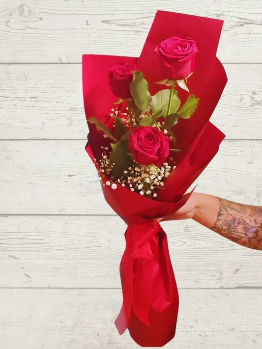A hand holding a gift-wrapped bouquet of red roses with green foliage and a red ribbon.