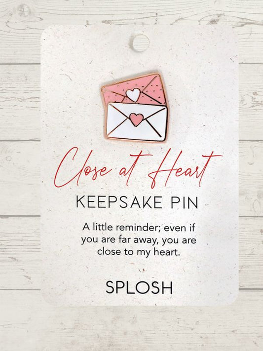 A keepsake pin with a ceramic heart-shaped mailbox design and a rose gold background, accompanied by an inspirational quote.