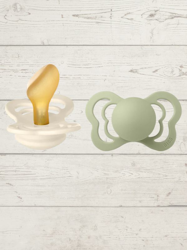 Two BIBS pacifiers with an anatomical glow design, one pacifier with a green ring on the shield and the other with a black ring, placed on a textured surface.