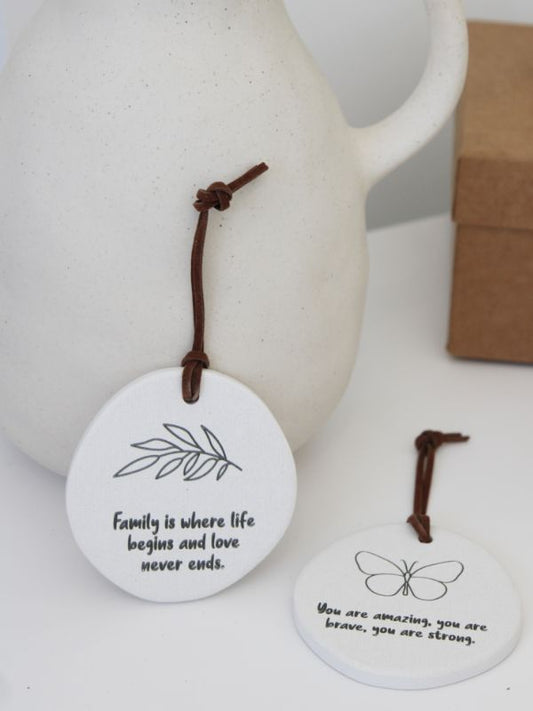 A white ceramic plaque with embossed text and a rope for hanging, displayed in front of a brown object which appears to be a box or bag.