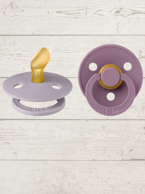 Two BIBS brand pacifiers, one in purple and one in blue, each with a round shield and a ring handle. The purple pacifier has a gold-colored nipple, and both display the BIBS logo.