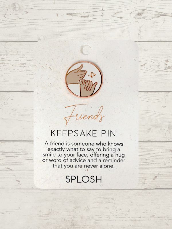 A keepsake pin with the word 'Friends' written on it, along with a heart symbol, in rose gold color, displayed against a white wooden background.