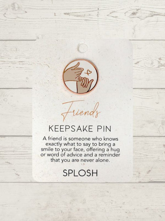 A keepsake pin with the word 'Friends' written on it, along with a heart symbol, in rose gold color, displayed against a white wooden background.