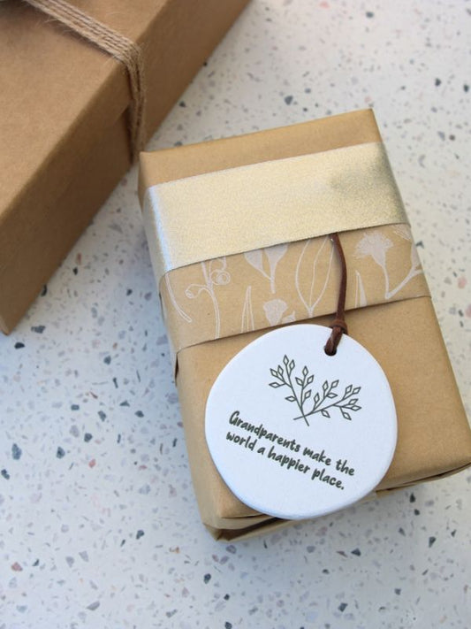 A beige ceramic plaque with embossed text and floral design, tied with a string and presented in a brown cardboard box with a rope handle.