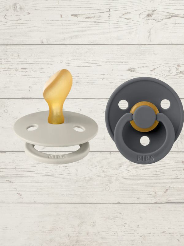 Two BIBS pacifiers with an anatomical glow design, one pacifier with a green ring on the shield and the other with a black ring, placed on a textured surface.