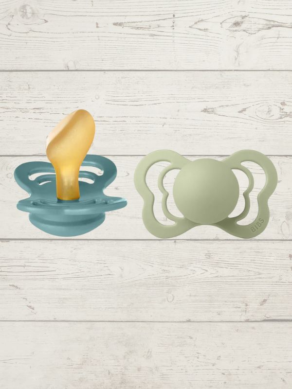 Two BIBS brand pacifiers, one with a yellow nipple and one with a green nipple, placed on a wooden surface.