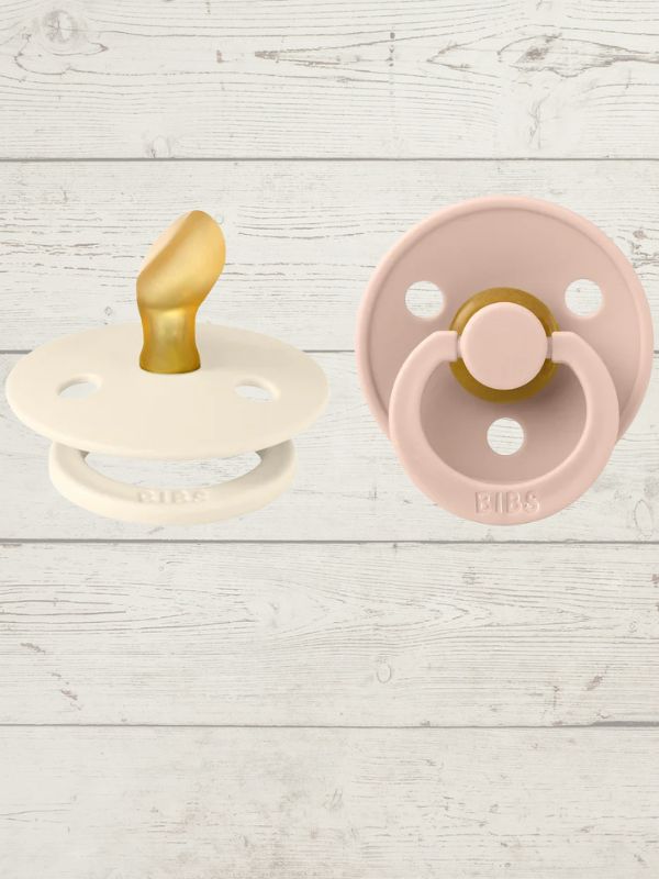 Two BIBS pacifiers with an anatomical glow design, one pacifier with a green ring on the shield and the other with a black ring, placed on a textured surface.
