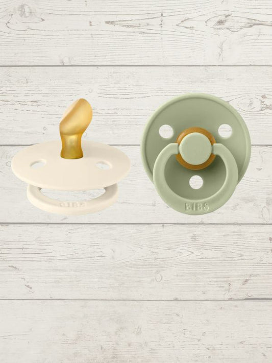 Two BIBS pacifiers with an anatomical glow design, one pacifier with a green ring on the shield and the other with a black ring, placed on a textured surface.