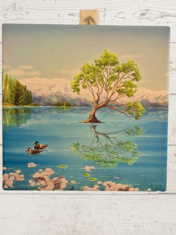 A ceramic tile featuring a scenic view of Lake Wanaka with a tree in the foreground, a cloudy sky, and a pair of ducks on the water.