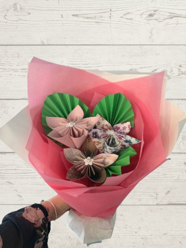 A bouquet of paper flowers with a mix of pink and green shades, arranged in a unique origami-style design, held against a wooden background.