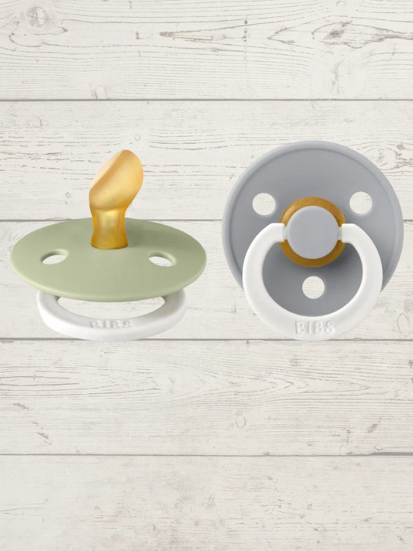 Two BIBS pacifiers with an anatomical glow design, one pacifier with a green ring on the shield and the other with a black ring, placed on a textured surface.