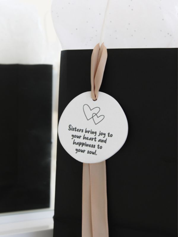 A black gift bag with a white and pink embossed ceramic plaque that reads 'Sisters bring joy to your heart and happiness to your soul.' It has a rope for hanging.