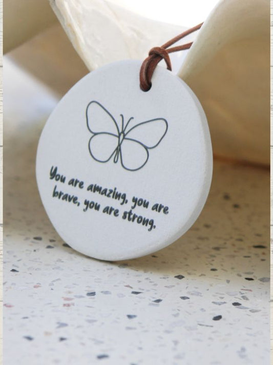 A white ceramic plaque with a butterfly design and the text 'You are amazing, you are brave, you are strong.' embossed on it, accompanied by a brown rope for hanging.