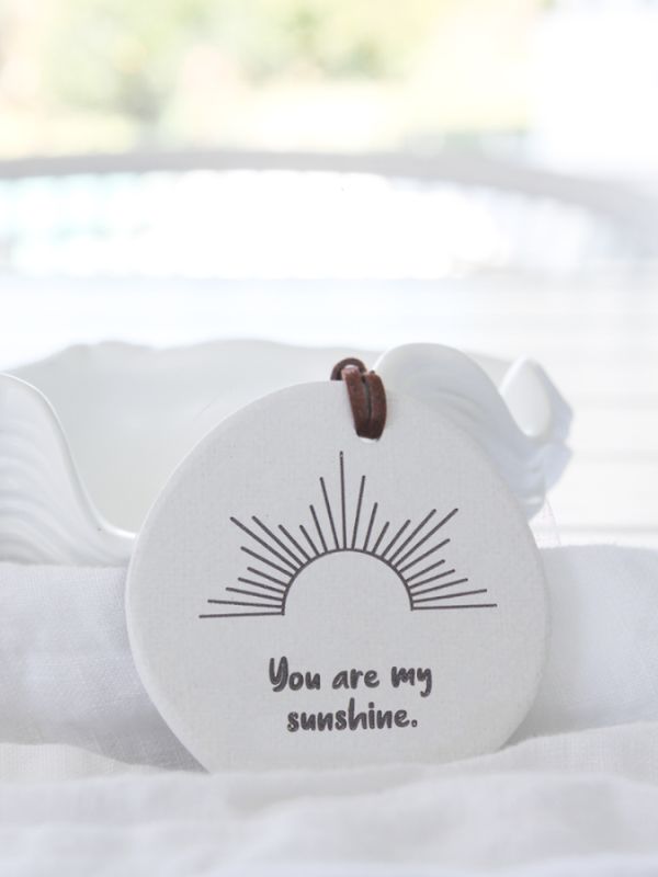 A white ceramic plaque with the text 'You are my sunshine' and a graphic of a sun and rain on it, with a brown rope for hanging.