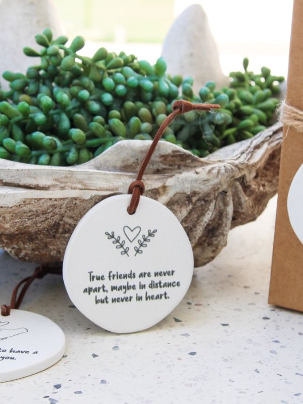A white ceramic plaque with embossed text and heartfelt quote, hanging by a rope next to a green plant in a rustic bowl.
