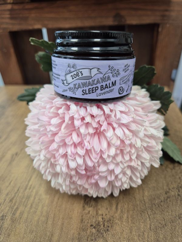A jar of Zoe's Kawakawa Lavender Sleep Balm placed on a pink floral decoration, with the product label visible.