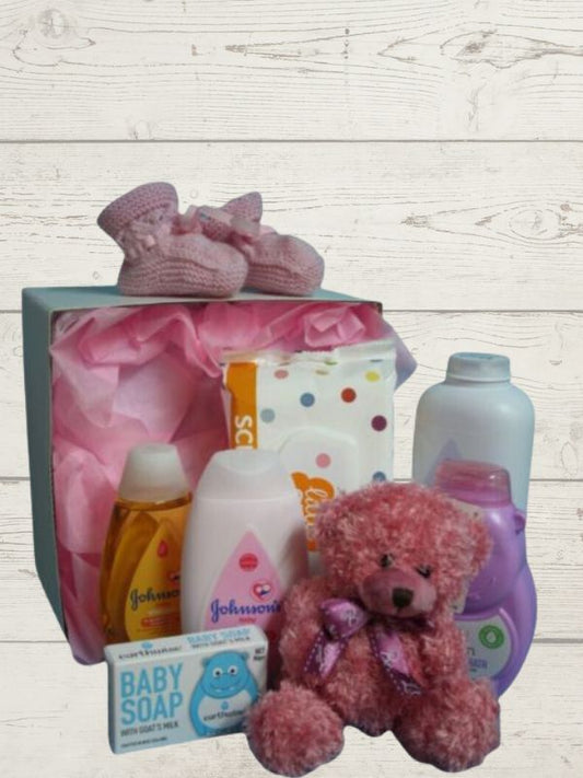A pink baby gift box opened to display a variety of baby care products including lotions, body wash, shampoo, wipes, a soft toy, and handmade booties.