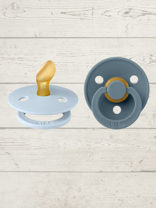 Two BIBS pacifiers with an anatomical glow design, one pacifier with a green ring on the shield and the other with a black ring, placed on a textured surface.