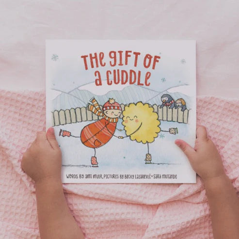 Child's hands holding a paperback book titled 'THE GIFT OF A CUDDLE' with colorful illustrations on the cover.