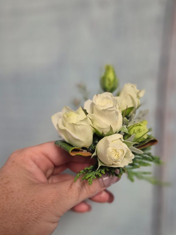 A hand holding a buttonhole made with white roses and green foliage.