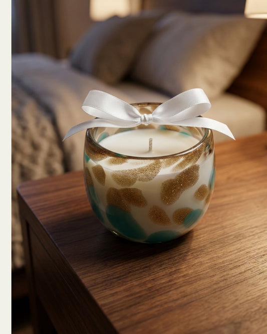 A decorative candle in a clear glass jar sits on a wooden bedside table in a softly lit bedroom. The candle has a white wax fill, a white ribbon tied in a bow on top, and a pattern of gold glitter spots and teal blue rings.