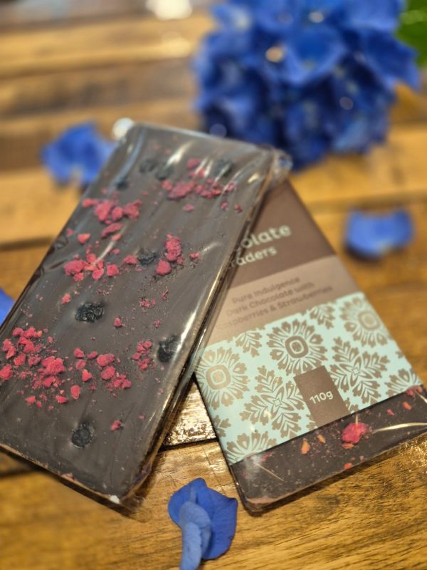 A chocolate bar with decorative toppings, placed on a wooden surface next to a flower, with the brand 'Chocolate Traders' visible on the packaging.