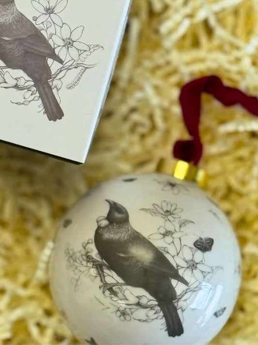 white bauble with a tui and flowers on it with a box on a brown background