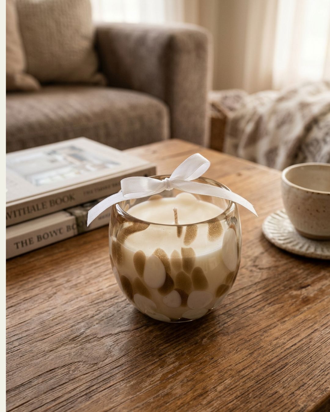 A glass jar candle with a white wax body, brown leaf-like swirls, and a white ribbon bow, sitting on a wooden table in a cozy living room setting.