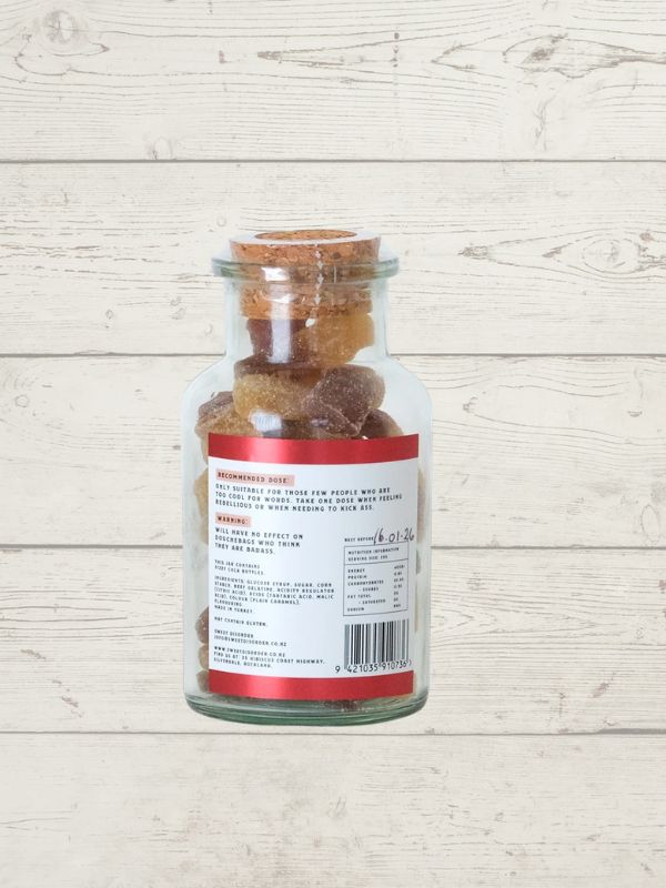 A clear glass jar with a cork stopper containing brown gummy candies, shown from the back with a white label featuring red borders and black text, set against a light wood plank background.