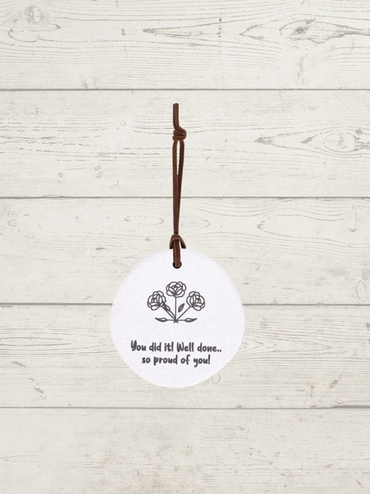 A white circular hanging ornament with a brown hanging cord, featuring a black line drawing of three roses and the printed message 'You did it! Well done.