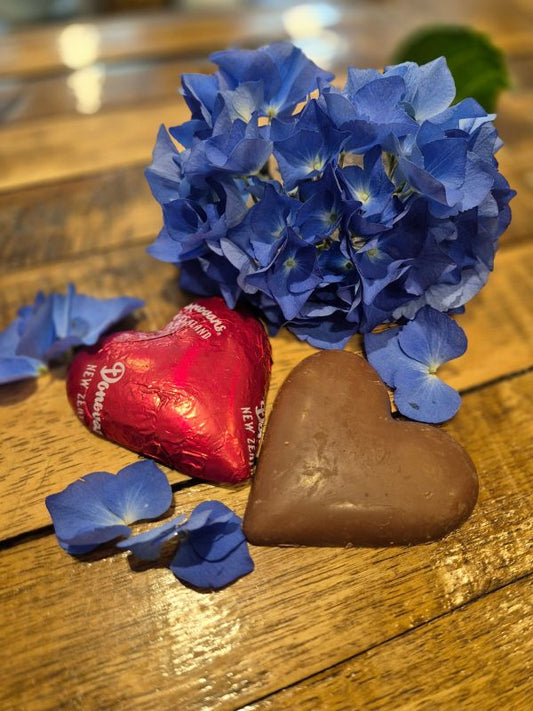A chocolate heart-shaped treat next to a blue flower on a wooden surface, with a red wrapper and a broken piece of chocolate visible in the background.