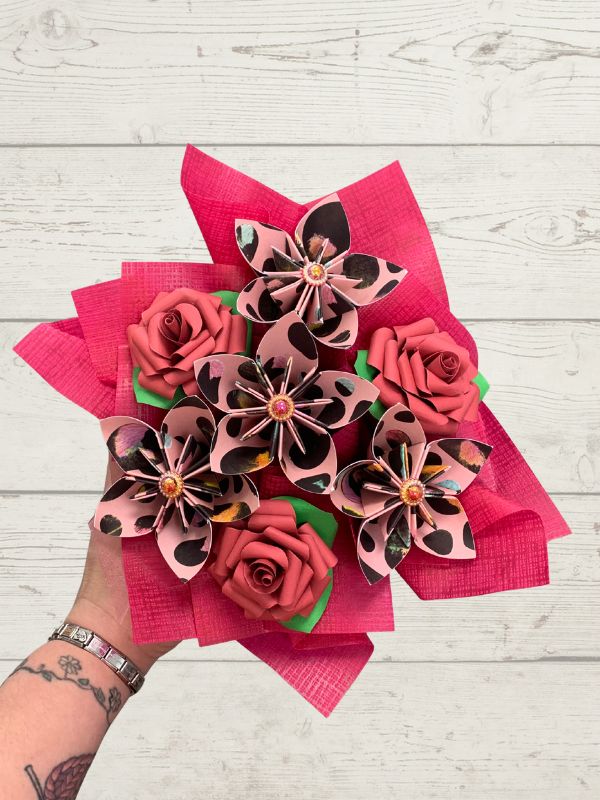 A hand holding a bouquet of artificial flowers made from pink patterned paper, including rose-shaped blooms and spotted petal flowers, wrapped in bright pink tissue paper, against a white wood plank background.