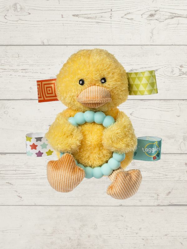 A yellow plush duck toy with a light blue bead choker, set against a white wood plank background. The duck has patterned fabric tabs on its head and body, and a small teal tag labeled 'taggies' is visible on its right side.