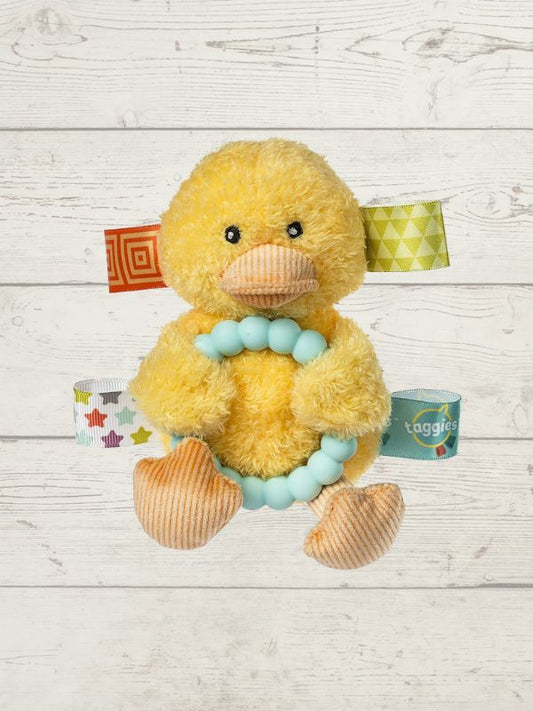 A yellow plush duck toy with a light blue bead choker, set against a white wood plank background. The duck has patterned fabric tabs on its head and body, and a small teal tag labeled 'taggies' is visible on its right side.