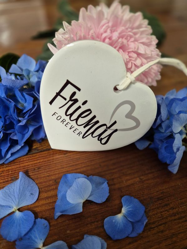 A white ceramic heart with the inscription 'Friends Forever' and a pink flower attached to it, placed on a wooden surface with blue flower petals scattered around.