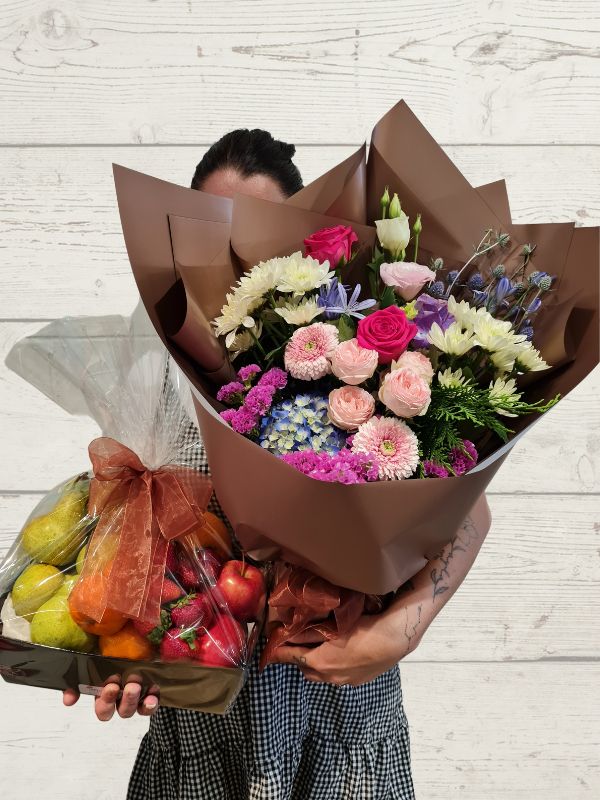 gift box of fruit and a bouquet of flowers held by a person on a white wooden background