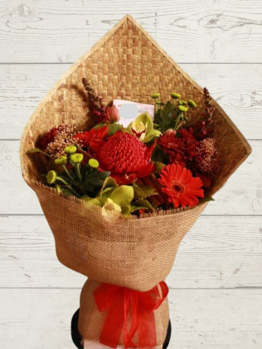 A fresh bouquet of red flowers wrapped in a harakeke mat, standing in a wooden vase on a wooden surface.