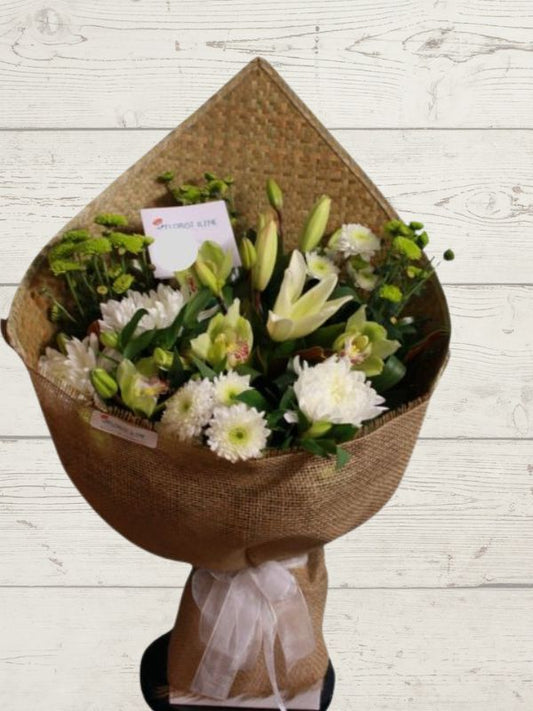 A bouquet of white and green flowers wrapped in a Harakeke mat with hessian trimmings, standing against a wooden background.