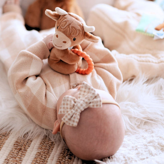 Baby wearing a checkered bow tie with a soft toy on a fluffy blanket with a highland cow rattle