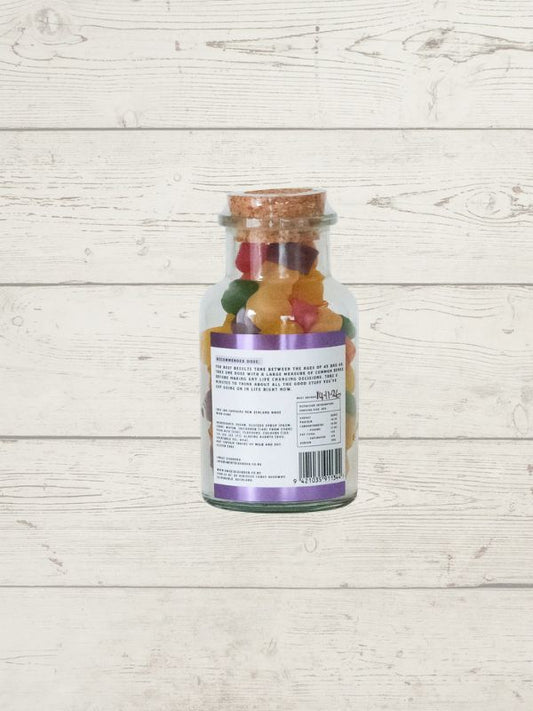 A clear glass jar filled with colorful gummy candies, sealed with a cork stopper and shown from the back with a white label, set against a light whitewashed wood plank background.