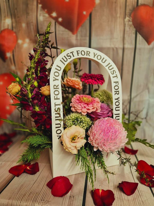 A colorful floral arrangement with a variety of flowers and greenery, displayed in a container with the text 'JUST FOR YOU' on it, against a wooden backdrop with heart decorations.