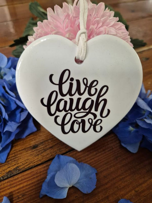 A white ceramic heart-shaped plaque with the words 'Live Laugh Love' written in black script, displayed on a wooden surface surrounded by blue flowers and pink-flowering plants.