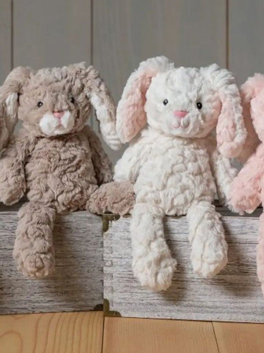Two soft toy bunnies with beige and white coloring, sitting on a wooden surface. One bunny is plush and the other is a stuffed toy, both with embroidered eyes and a textured surface.