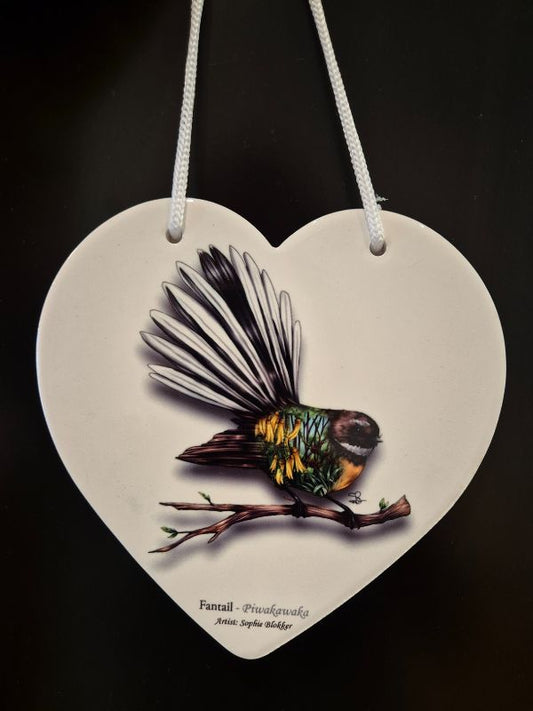 A ceramic heart-shaped plaque with a Fantail bird artwork on it, primarily in gray and beige tones, with a rope hanging at the top.