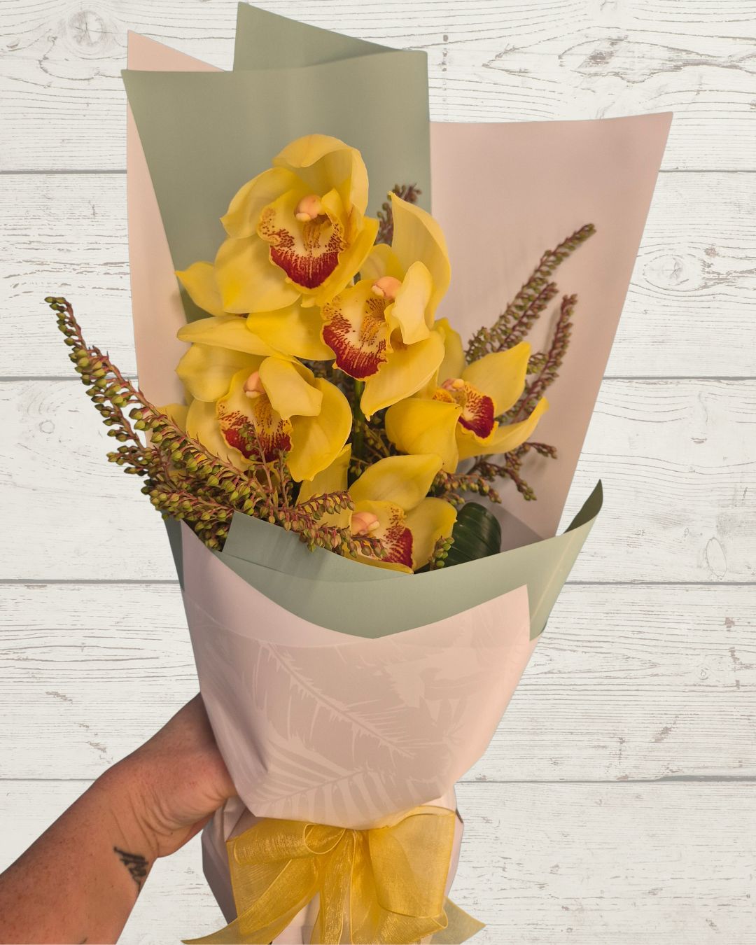 Bouquet of yellow flowers with green wrap held by a hand on a light wooden surface, Fresh flowers delivered in Hamilton