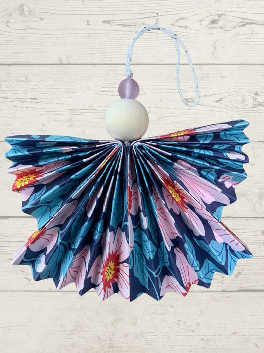 A decorative hanging ornament shaped like a stylized angel, featuring a pleated fabric body with a blue, pink, and teal floral pattern, a white wooden bead, a small lavender bead, and a light blue hanging string against a whitewashed wood background.