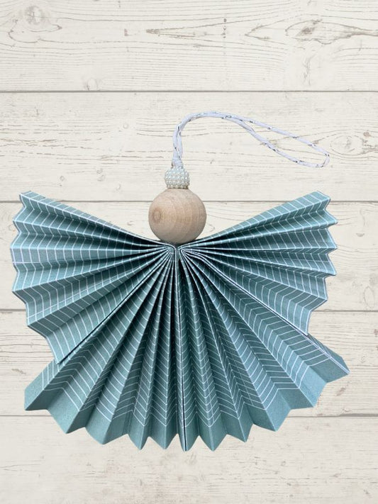 A decorative hanging ornament shaped like a paper angel, featuring a pleated blue and white striped wing, a round natural wood torso, and a white hanging string, displayed against a light whitewashed wood plank background.
