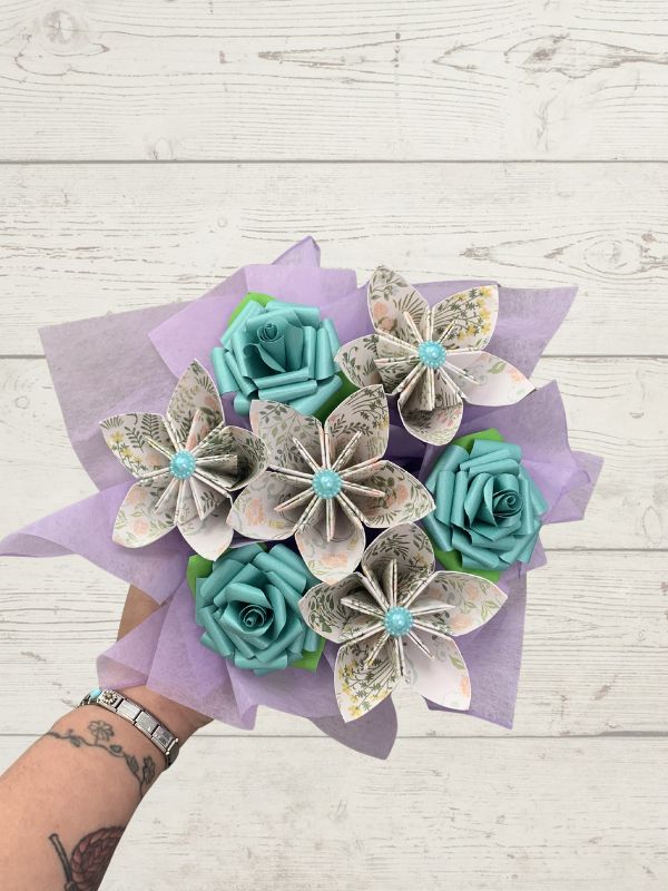 A hand holding a bouquet of paper flowers made from book pages, featuring teal paper roses and patterned paper flowers wrapped in light purple tissue paper, set against a white wood plank background.