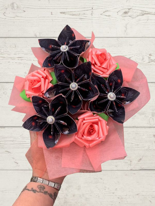 A hand holding a bouquet of artificial flowers, including black star-shaped blossoms with pearl-like centers and pink paper roses, wrapped in pink tissue paper against a white wood plank background.