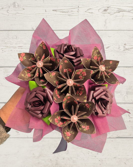 A bouquet of handmade origami paper flowers, featuring dark brown patterned flower petals with pink floral details and purple paper roses, wrapped in pink tissue paper and held by a hand against a white wood background.